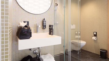 A modern bathroom with a glass-enclosed shower, a round mirror, a white sink, and a black handbag on the counter.
