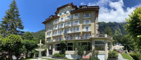 A multi-story hotel with balconies, a garden with a fountain, and a mountainous backdrop.