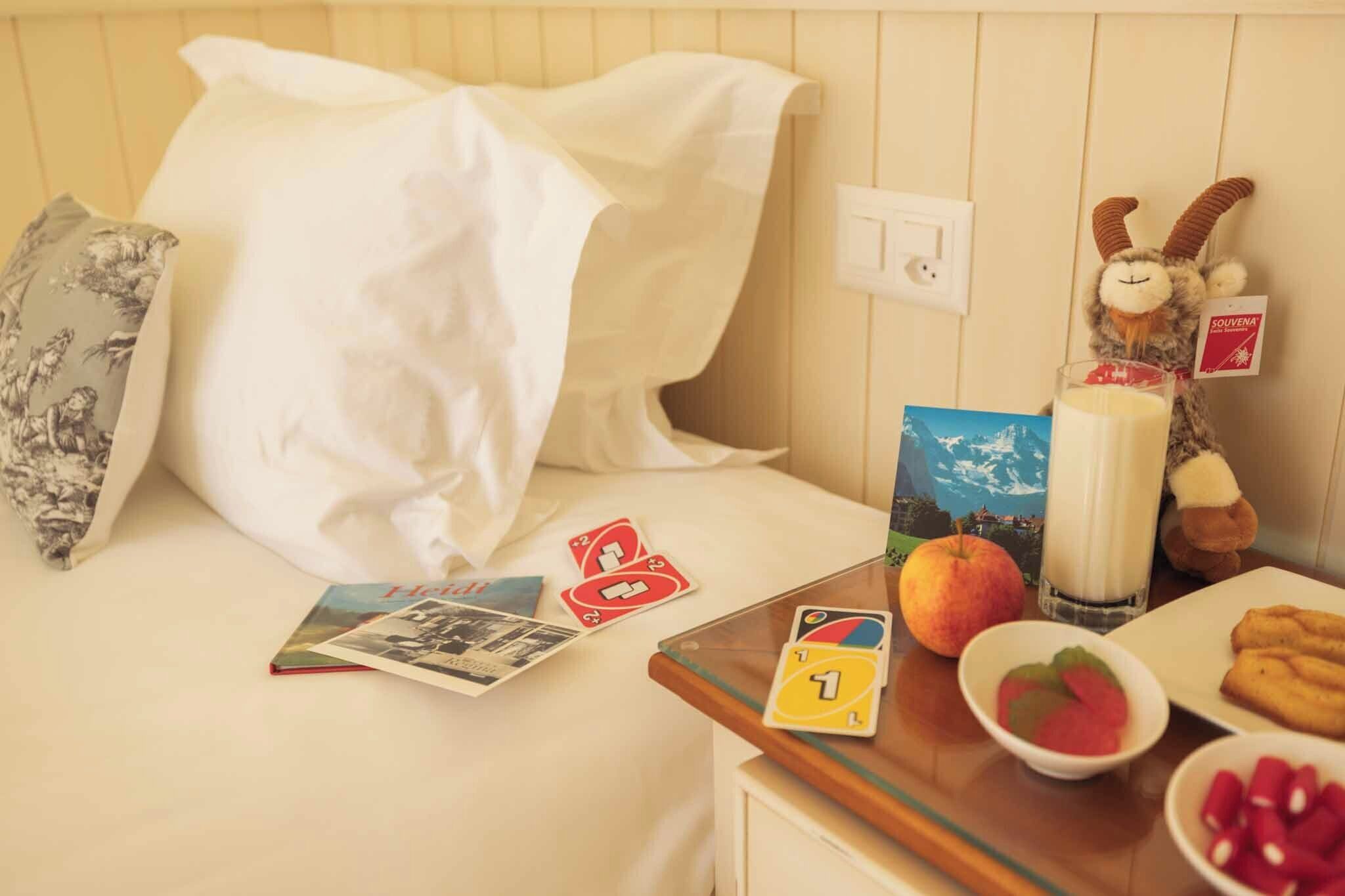 Un lit bien fait, avec du linge de lit blanc, un jouet en peluche, un verre de lait, un bol de fruits et une assiette de biscuits sur une petite table en bois.