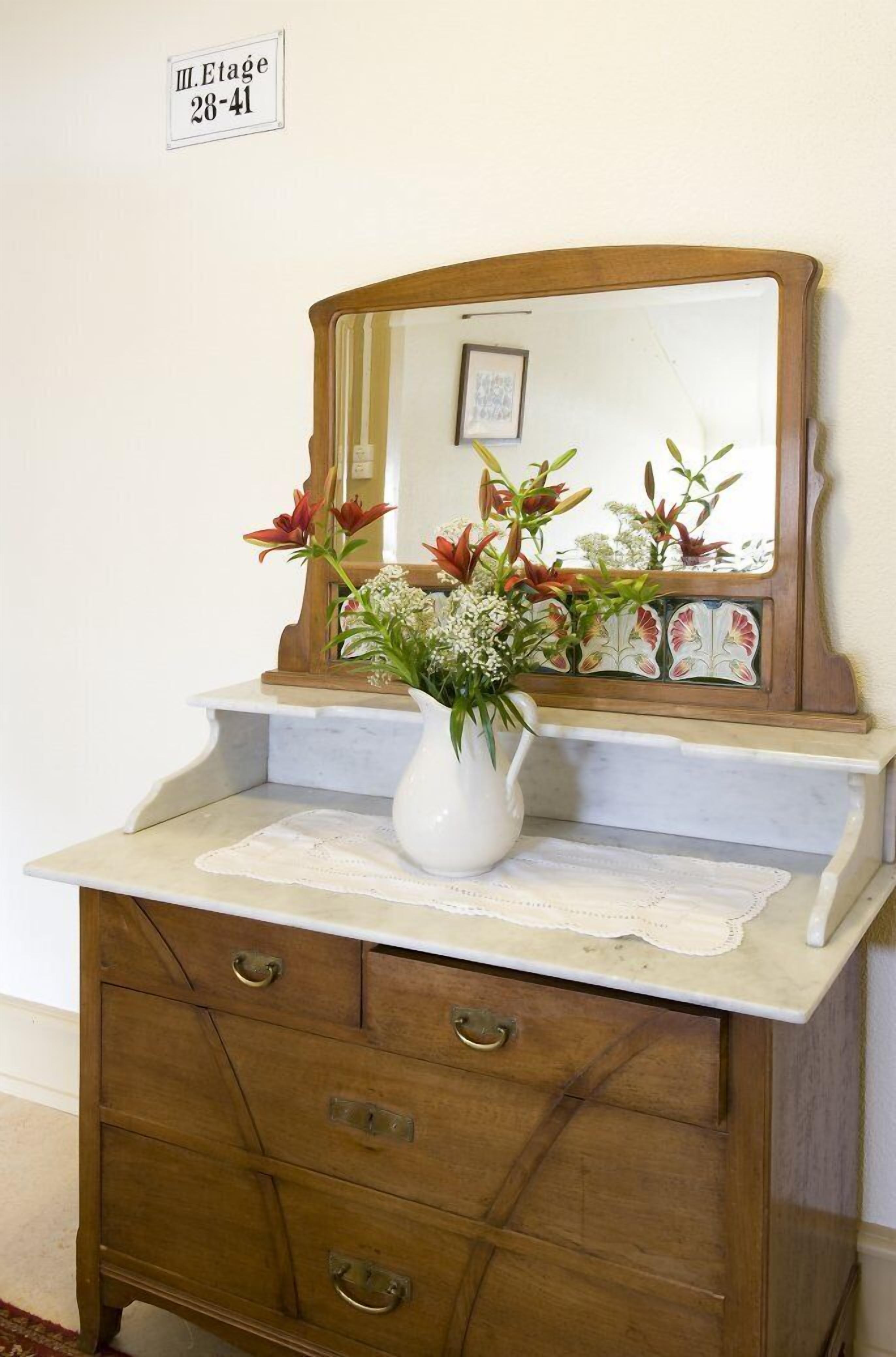Eine Holzkommode mit einer weißen Vase, die Blumen enthält, ein Spiegel darüber und eine Spitzenhäkelarbeit auf der Arbeitsfläche.