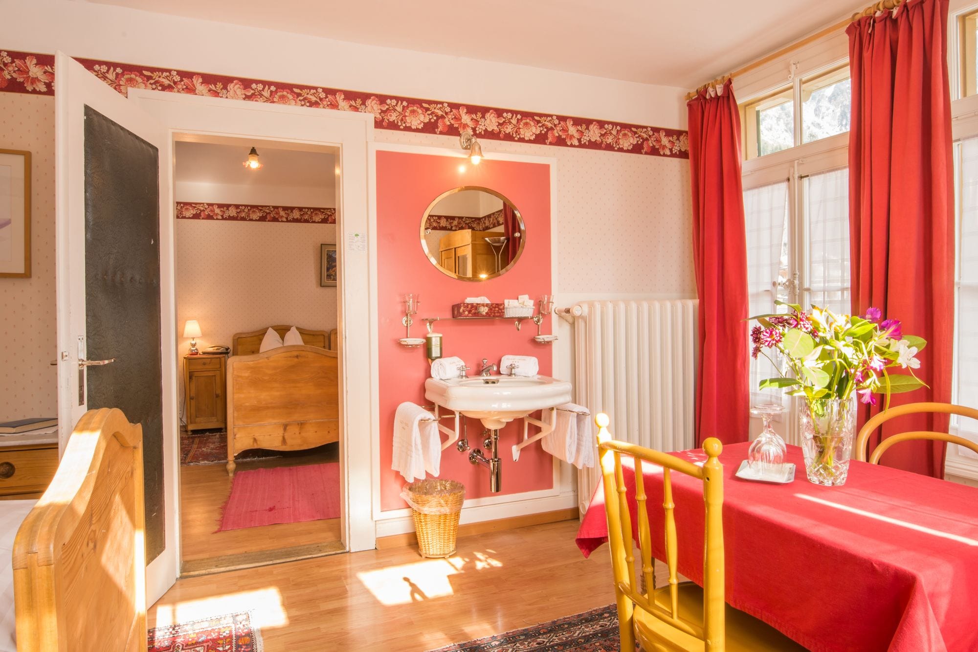 Una habitación con una cama de madera, una mesita de noche con una lámpara, un espejo con marco rojo, un lavamanos blanco, una mesa roja con un jarrón de flores y una ventana con cortinas rojas.