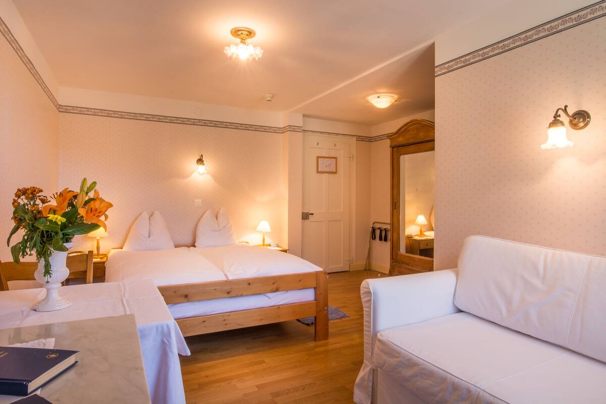 A hotel room with a wooden bed, a white sofa, a small table with a vase of flowers, and two bedside lamps.