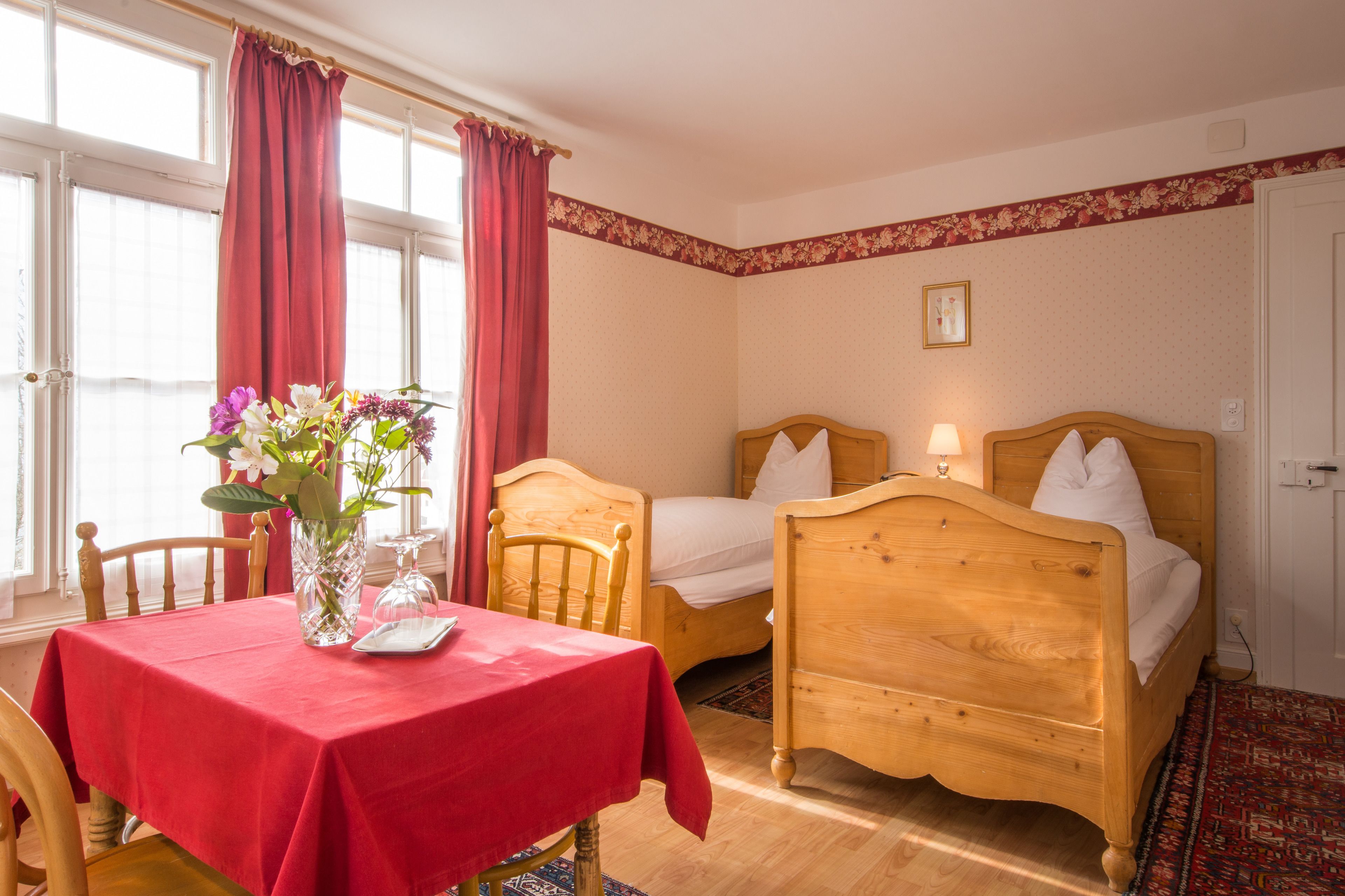 A room with two wooden beds, a dining table with a red tablecloth, a vase with flowers, and a window with red curtains.