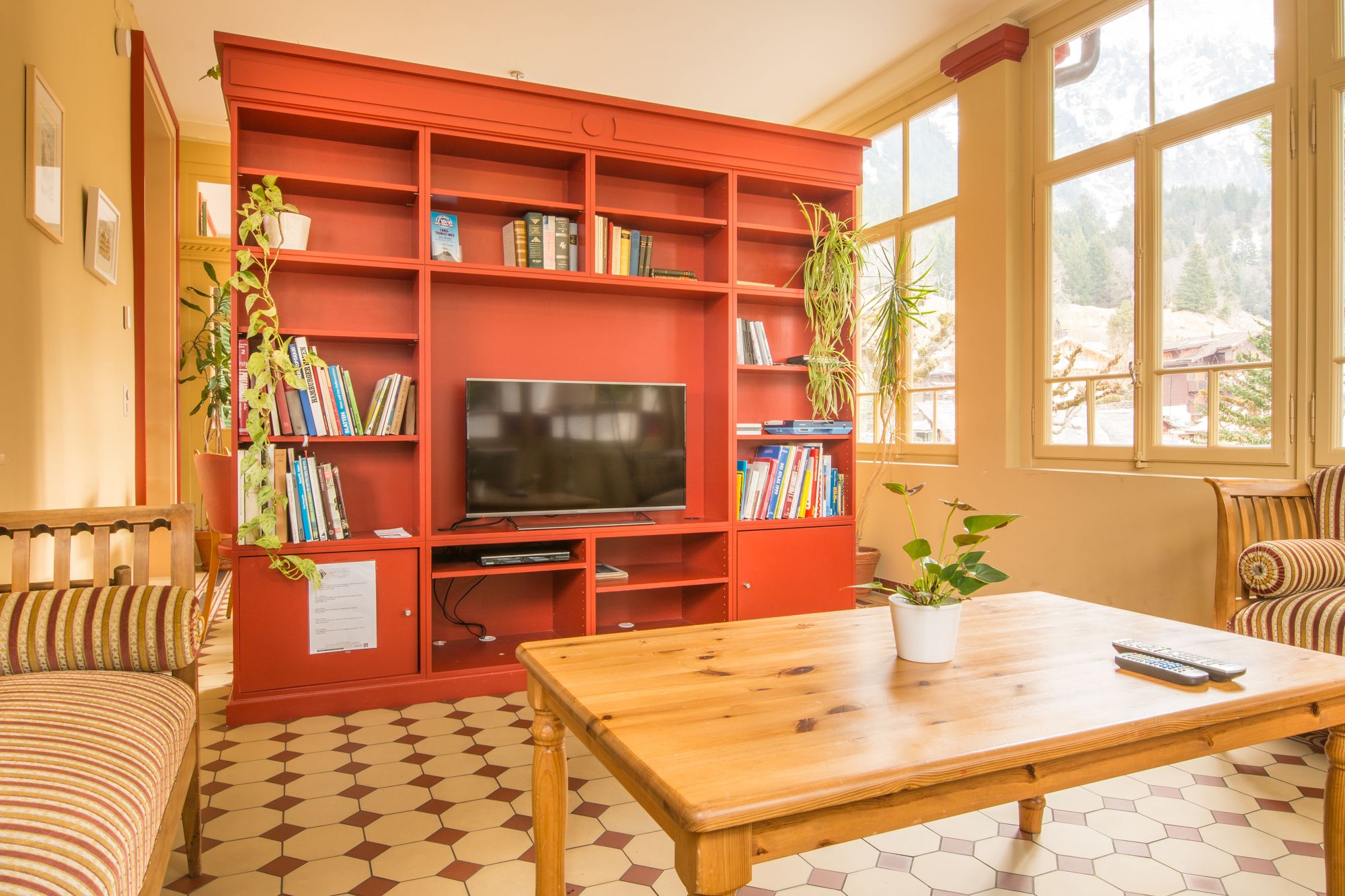 Ein gemütliches Wohnzimmer mit einem roten Bücherregal, einem Flachbildfernseher, einem Holztisch und zwei gestreiften Sofas.