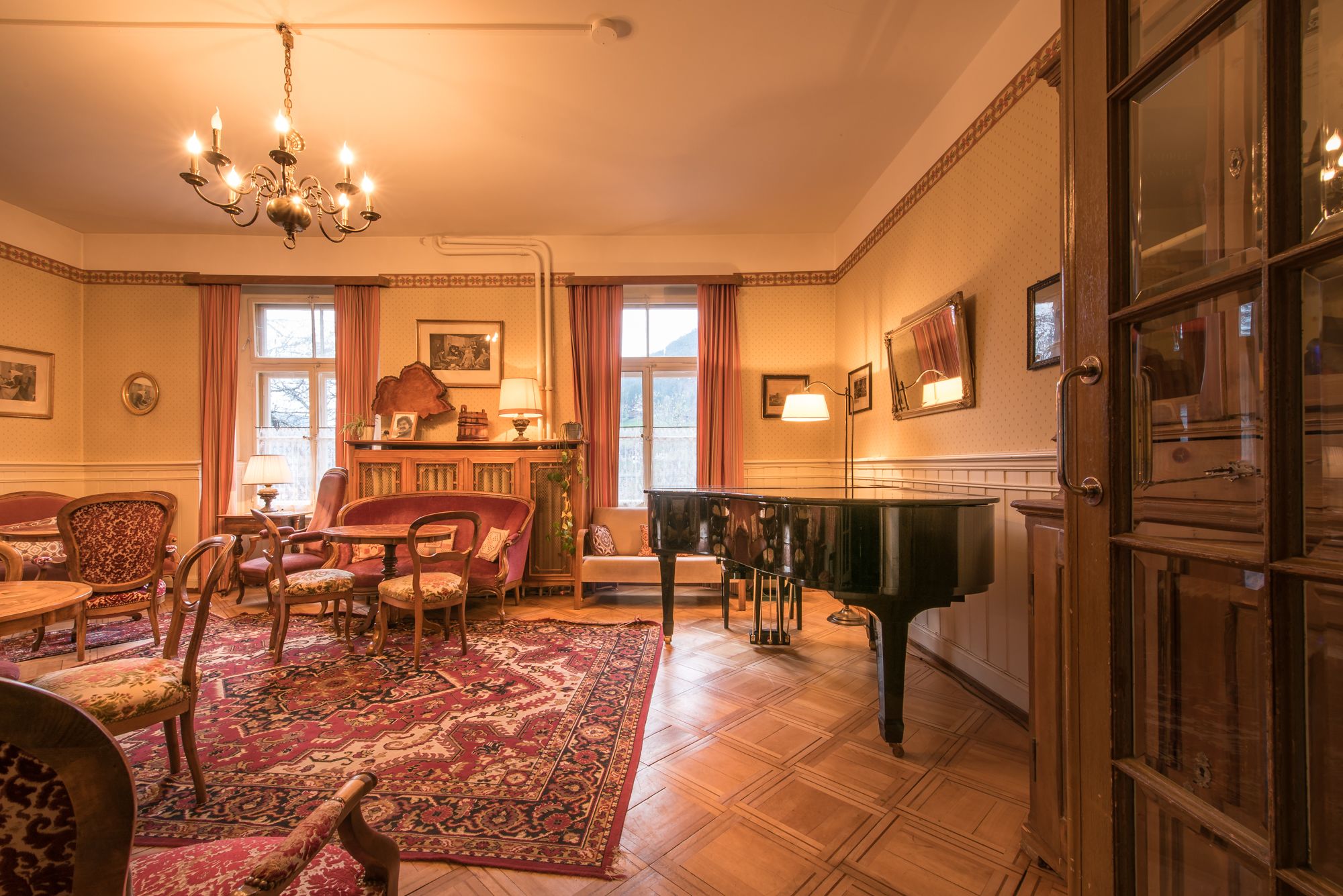 A spacious living room with a grand piano, a chandelier, and a large window with curtains.