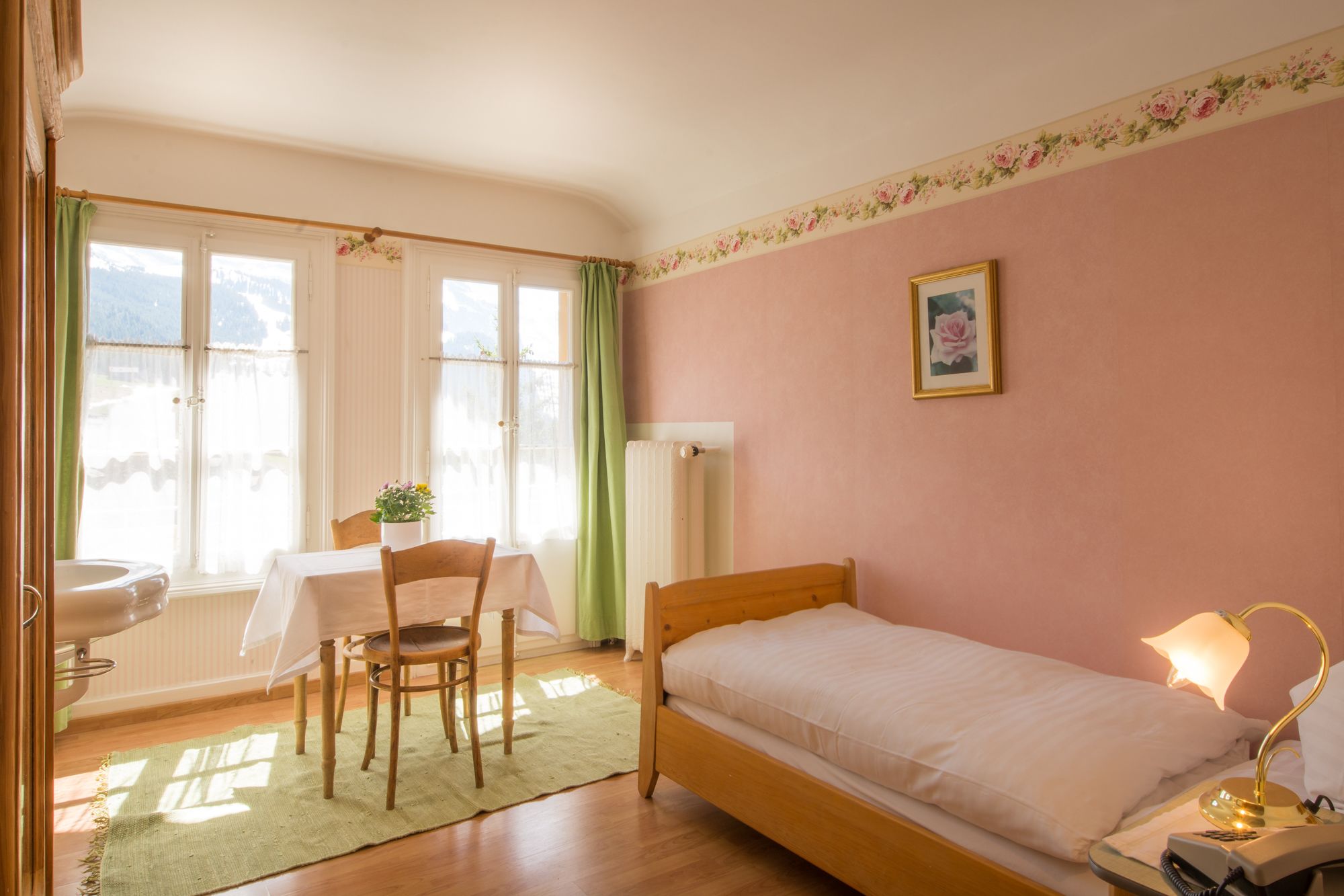 A bedroom with a single bed, a bedside table with a lamp, a chair, a table with a plant, and a window with curtains.