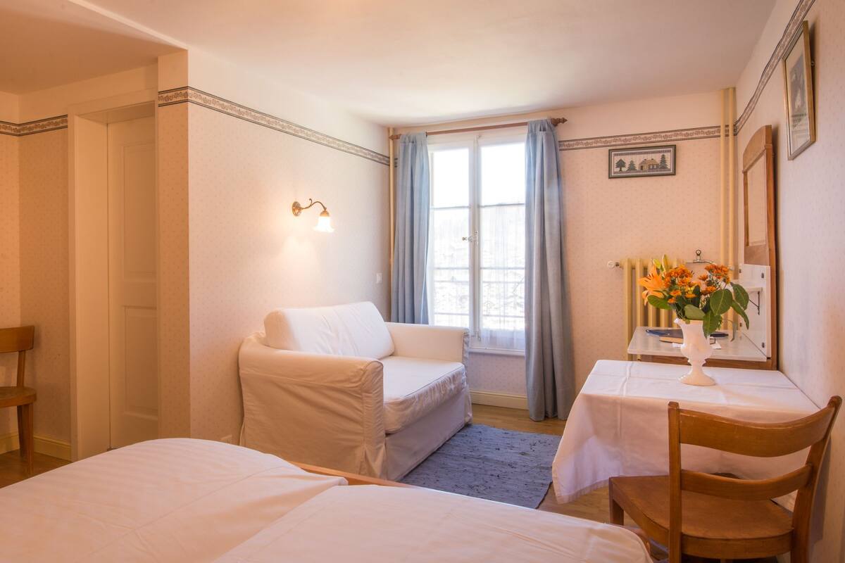 A hotel room with two single beds, a small table with a vase of flowers, a chair, and a window with curtains.