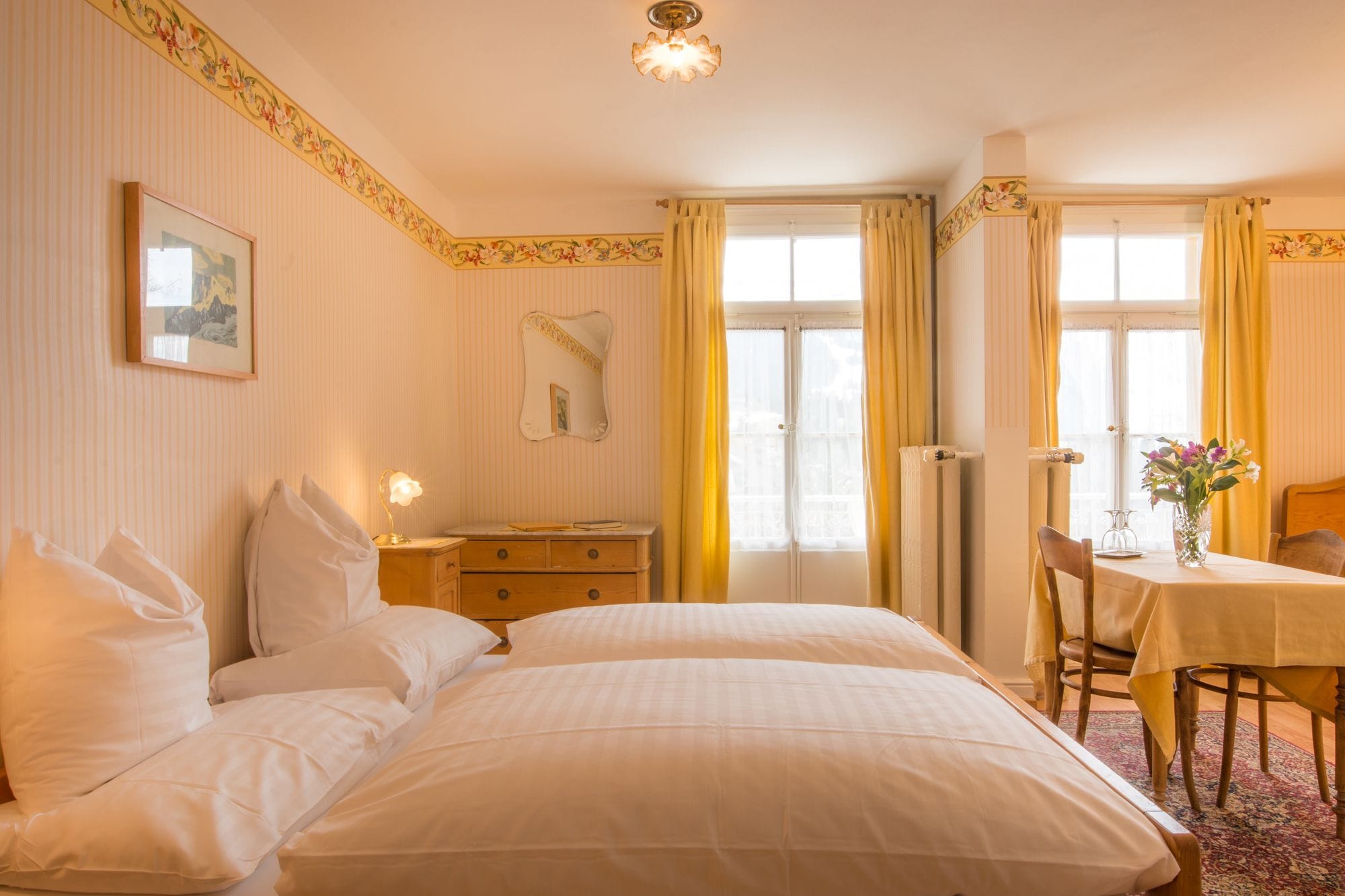 A hotel room with a large bed, a bedside table with a lamp, a chair, a small table with a vase, and a window with curtains.