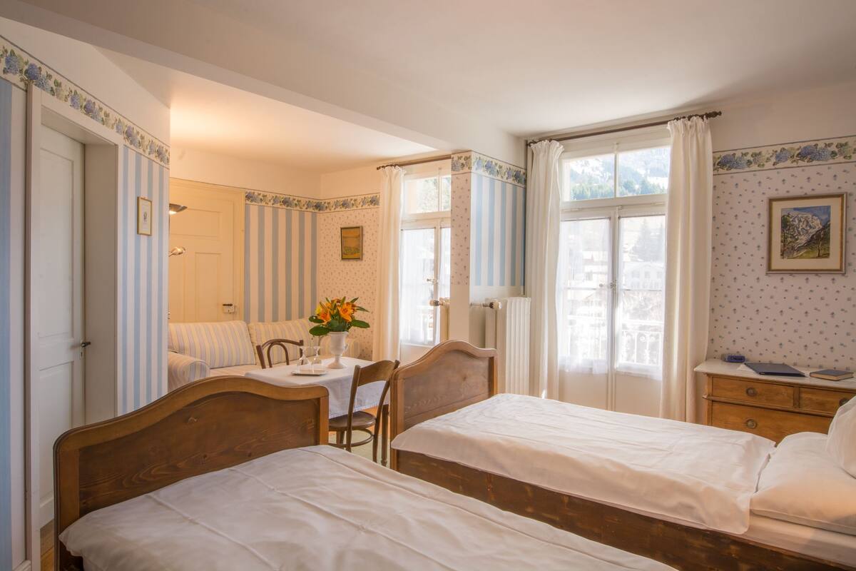 A bedroom with two single beds, a bedside table with a lamp, a chair, a small table with a vase of flowers, and a window with curtains.
