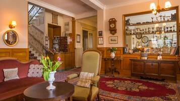 Un interior tradicional con un sofá, un sillón y una mesa redonda con un jarrón de flores. Se observa una escalera al fondo que conduce a otra habitación.