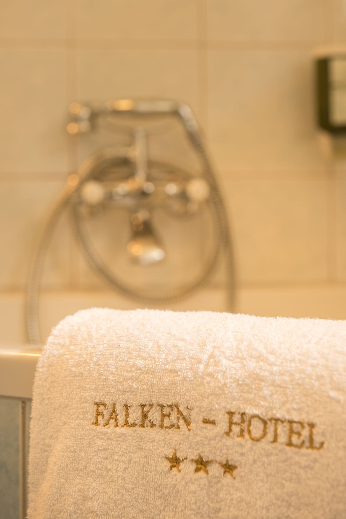 Una toalla blanca con el nombre "FALKEN-HOTEL" bordado en dorado, colgada en un baño con cabezal de ducha visible y paredes de azulejos.