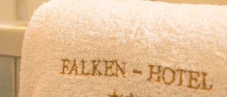 Una toalla blanca con el nombre "FALKEN-HOTEL" bordado en dorado, colgada en un baño con cabezal de ducha visible y paredes de azulejos.