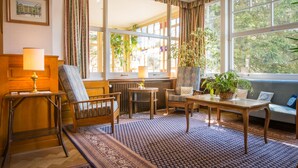 A cozy room with a wooden chair, a table, and a sofa. There are large windows with curtains, a lamp, and a framed picture on the wall. - Hotel Falken (Lauterbrunnen)