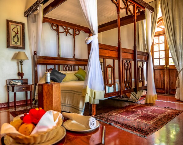 A four-poster bed with a canopy, a bedside table with a lamp, and a fruit basket on the floor.