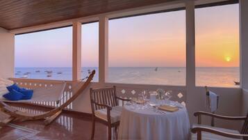 A room with a view of the sunset over the sea, a hammock, a table set for dining, and wooden chairs.