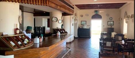 A bar area with a wooden counter and a person behind it. There are chairs and tables in the room.
