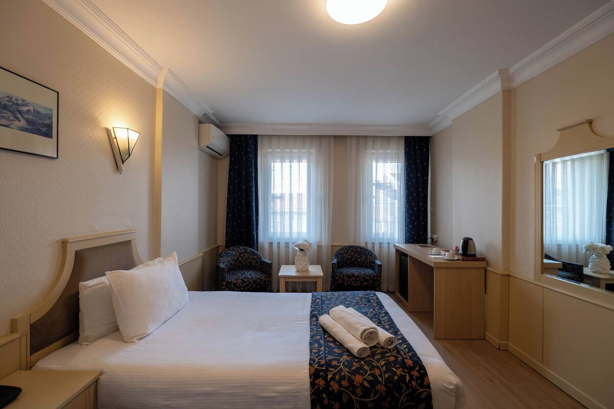 A hotel room with a bed, two armchairs, a small table with a vase, and a desk with a kettle.