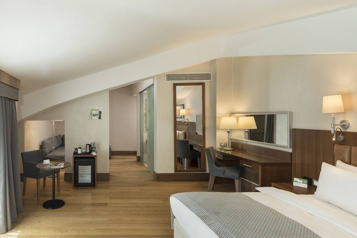 A hotel room with a bed, desk, chair, and a small table with a lamp.