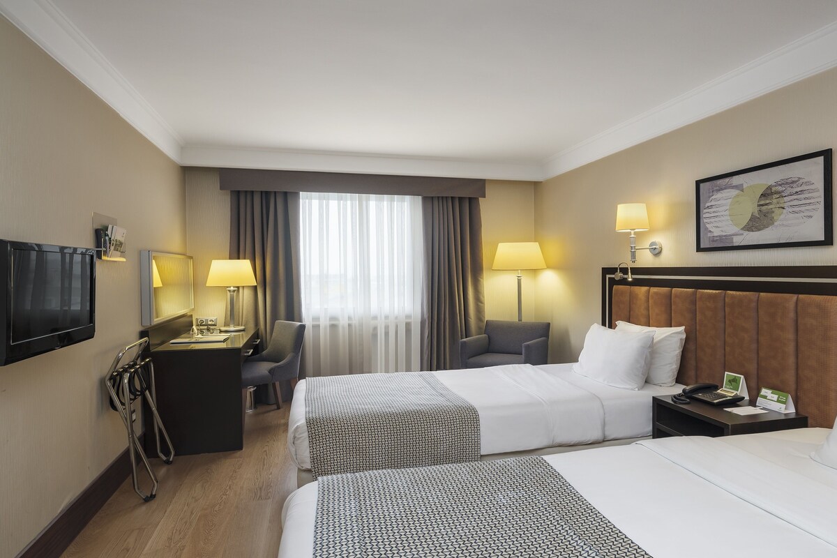 A hotel room with two beds, a desk with a lamp, a TV, and a window with curtains.