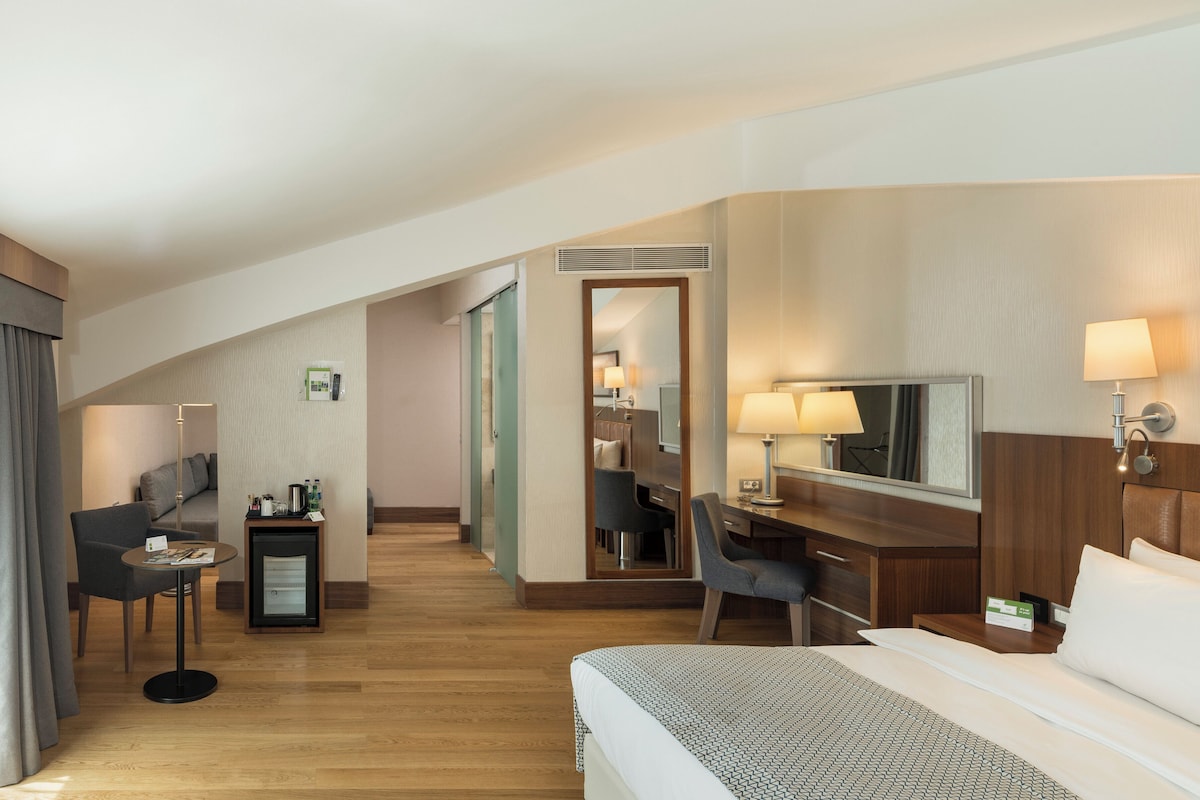 A hotel room with a bed, desk, chair, and a small table with a lamp.