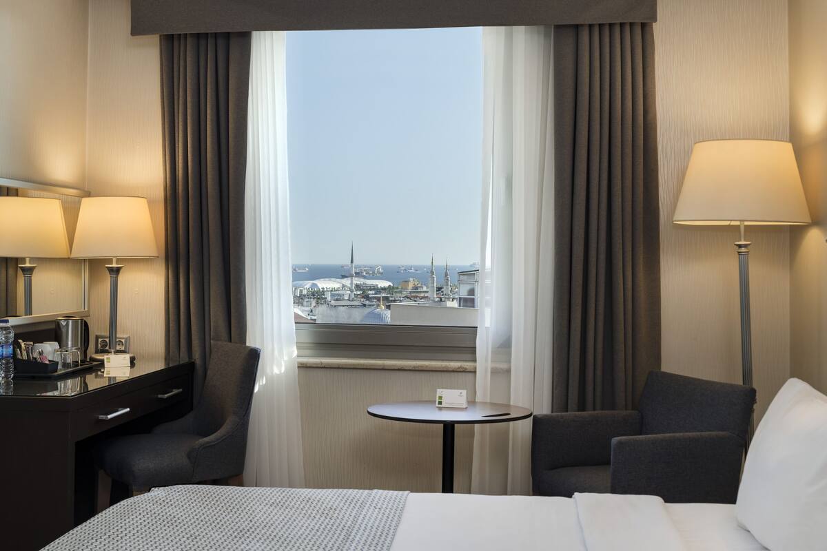 A hotel room with a bed, two armchairs, a small table, and a desk with a lamp and a coffee maker.