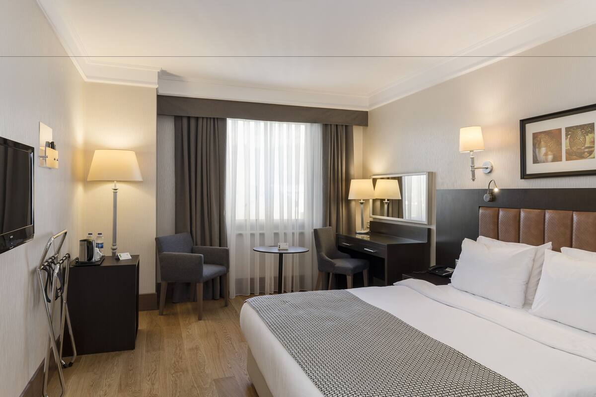 A hotel room with a large bed, a desk with a chair, a small table, a TV, and a window with curtains.
