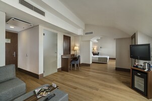 Habitación de hotel con piso de madera, un escritorio con silla, una cama, un televisor y una kitchenette pequeña.