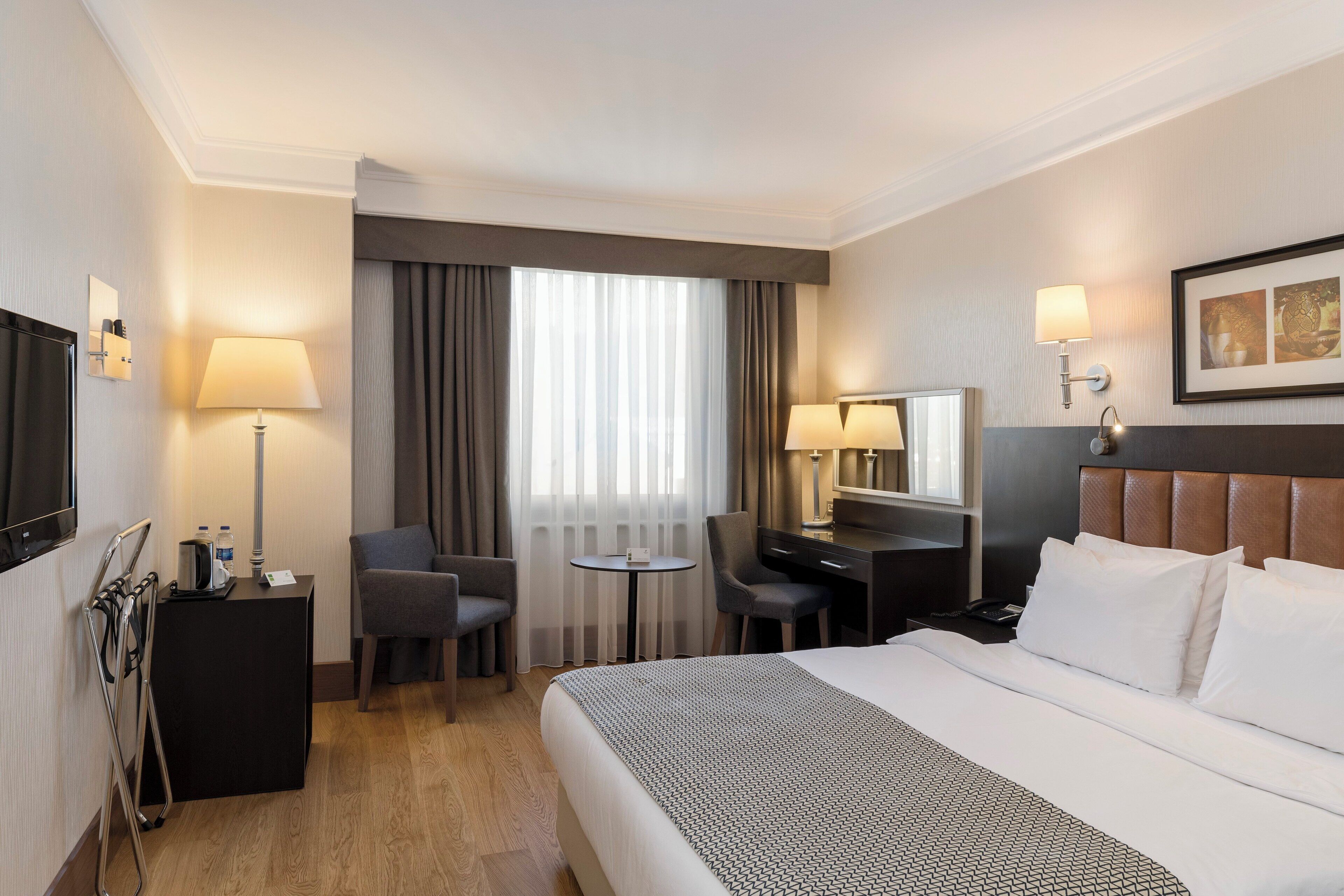 Une chambre d’hôtel comprenant un lit, deux tables de chevet, un bureau, une chaise, un téléviseur et une fenêtre avec des rideaux.