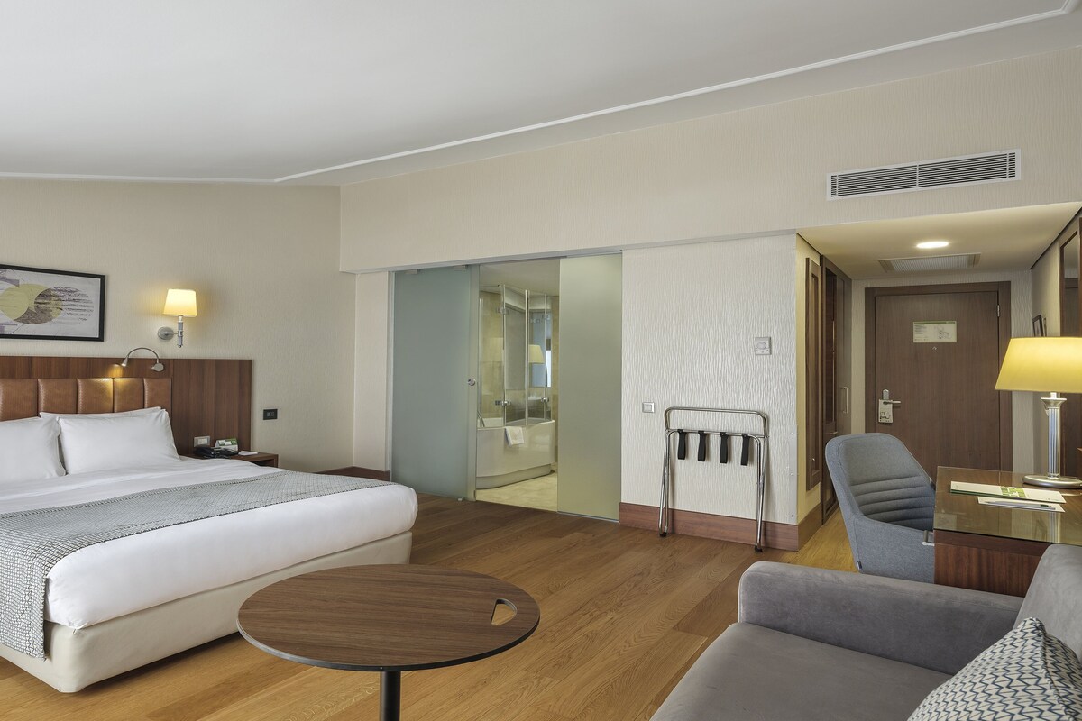A hotel room with a bed, bedside tables, a desk, a chair, a round table, a shower area, and a door.
