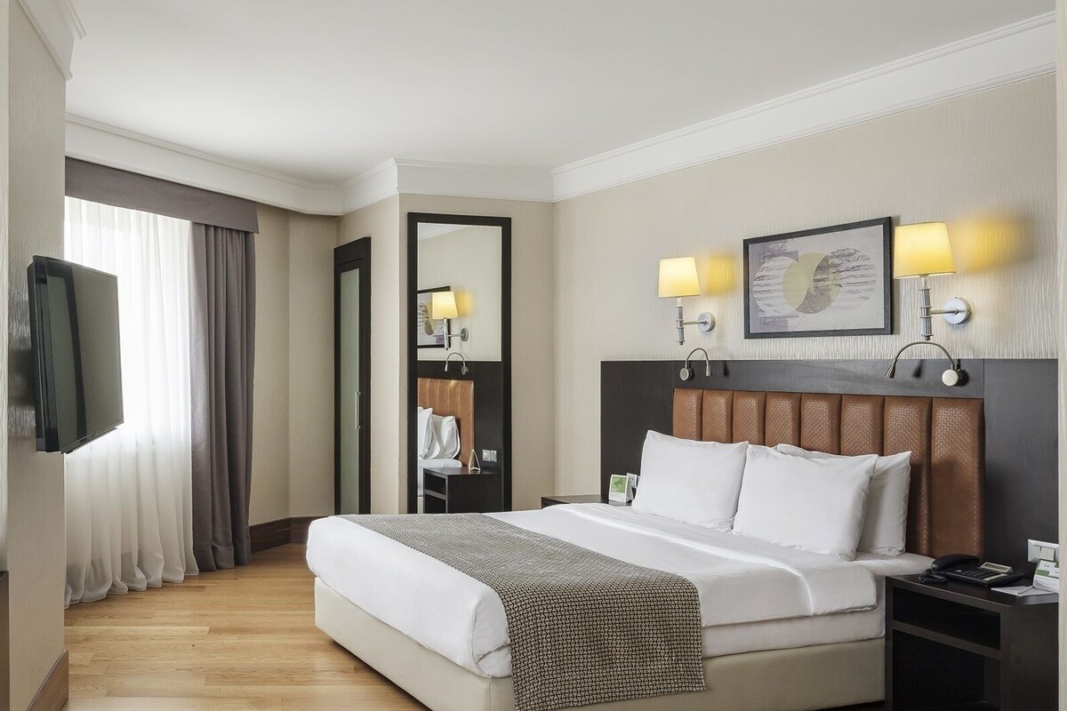 A hotel room with a large bed, bedside lamps, a flat-screen TV, a mirror, and a nightstand with a phone and a lamp.