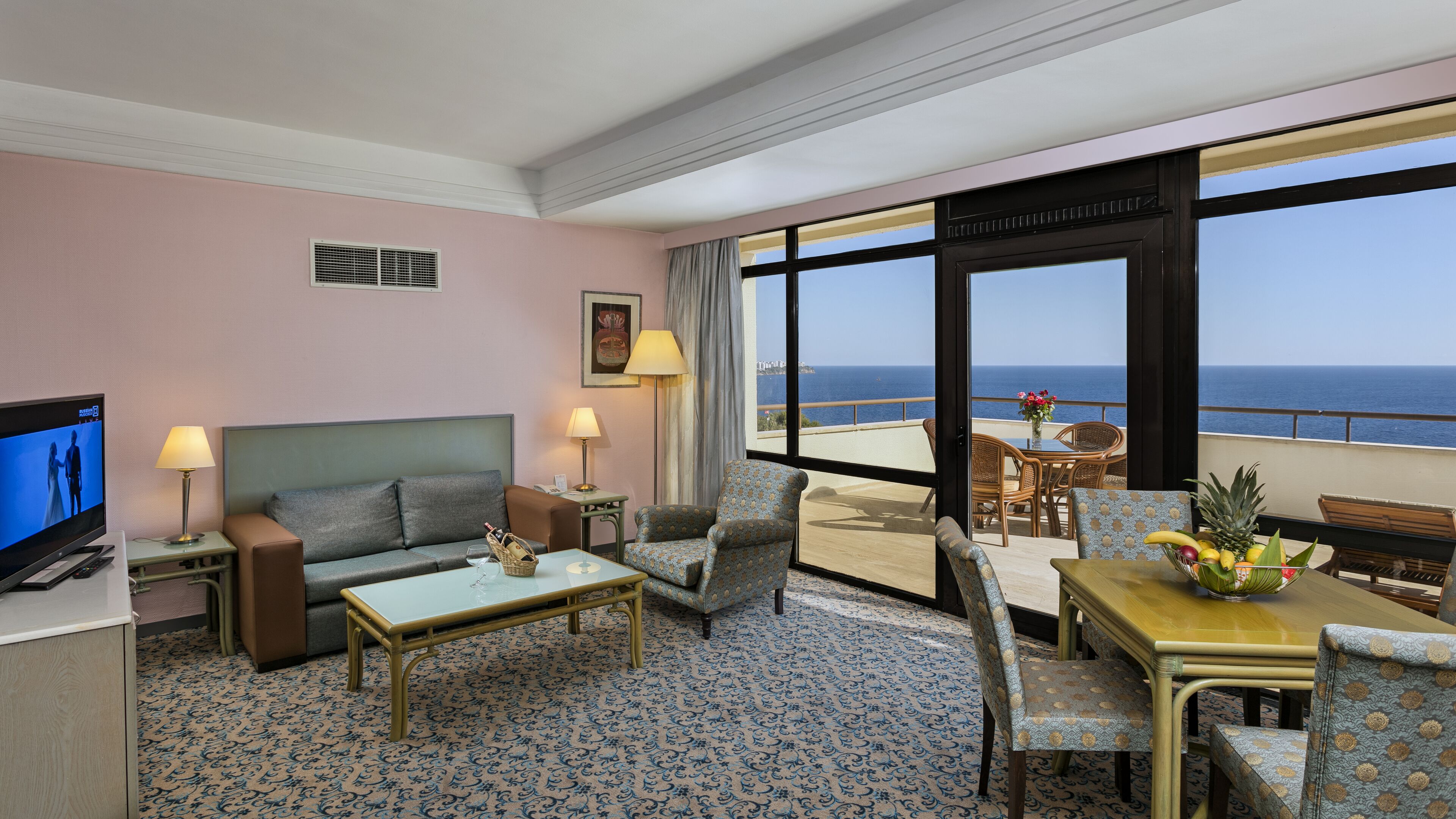 A hotel room with a sofa, armchair, coffee table, and a balcony with a table and chairs.