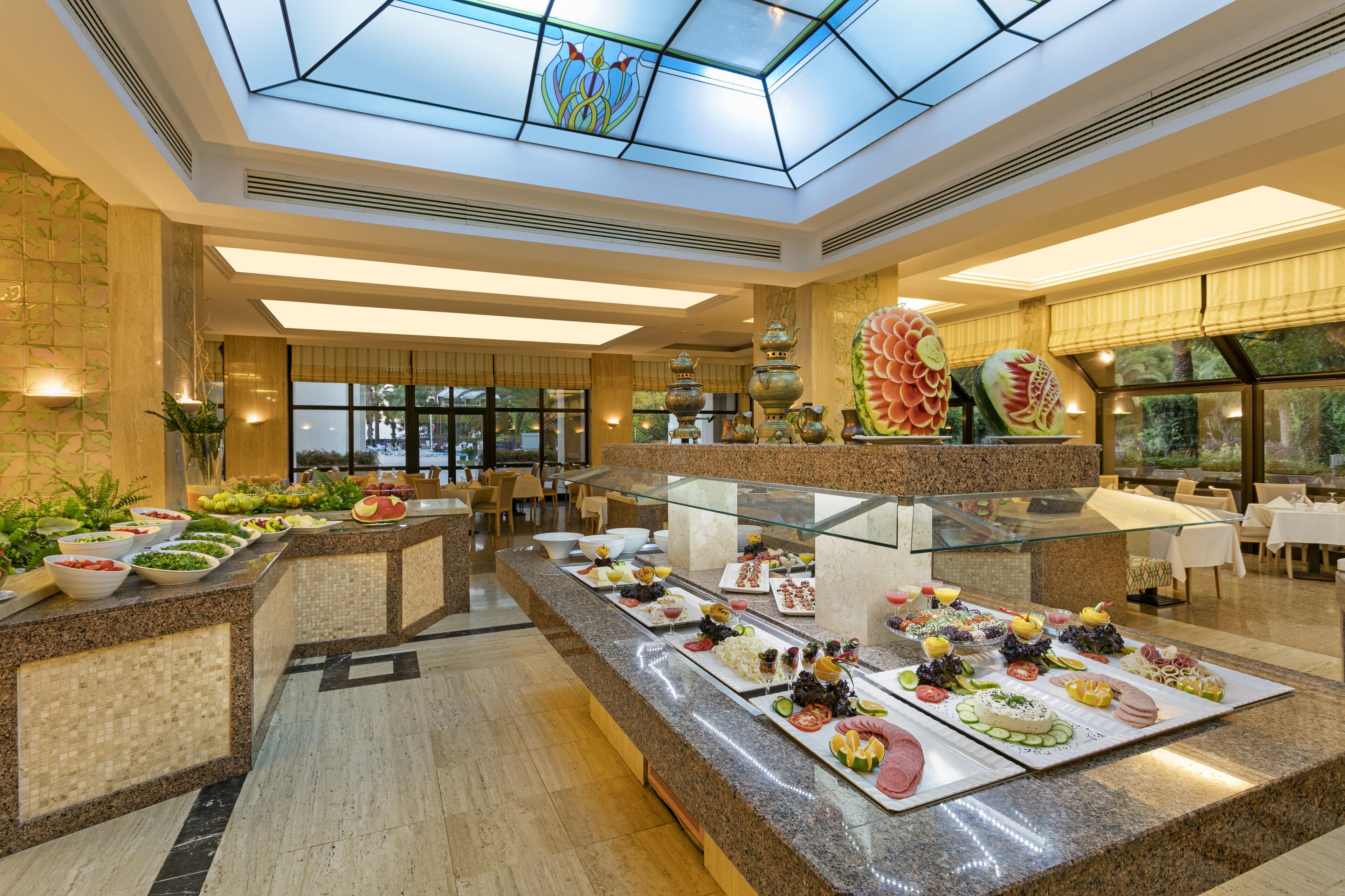A hotel dining area with a glass-top buffet counter, various food items, and a high ceiling with skylights.