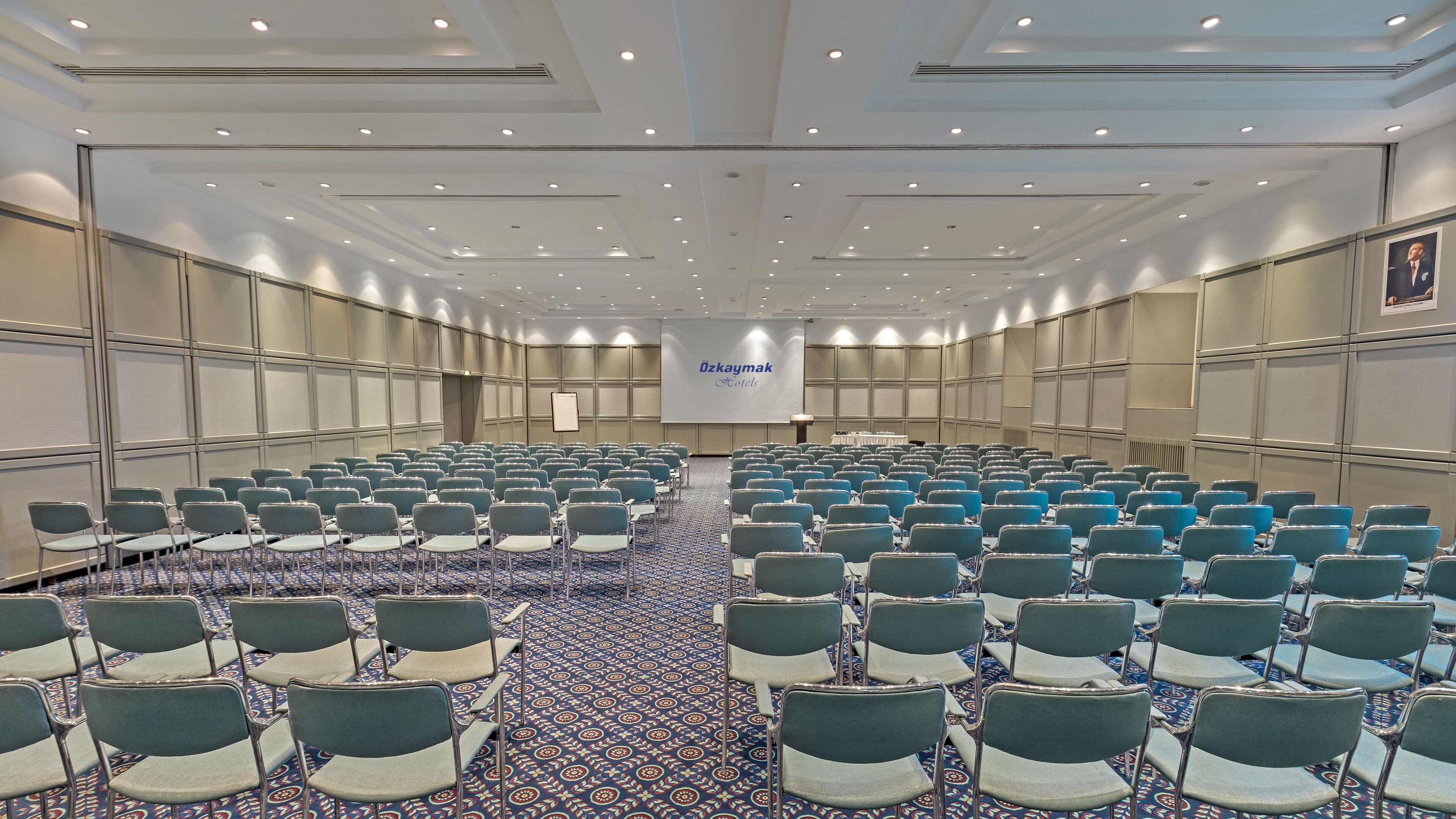 A large conference room with rows of chairs, a patterned carpet, and a banner reading "Özkaymak Hotels".