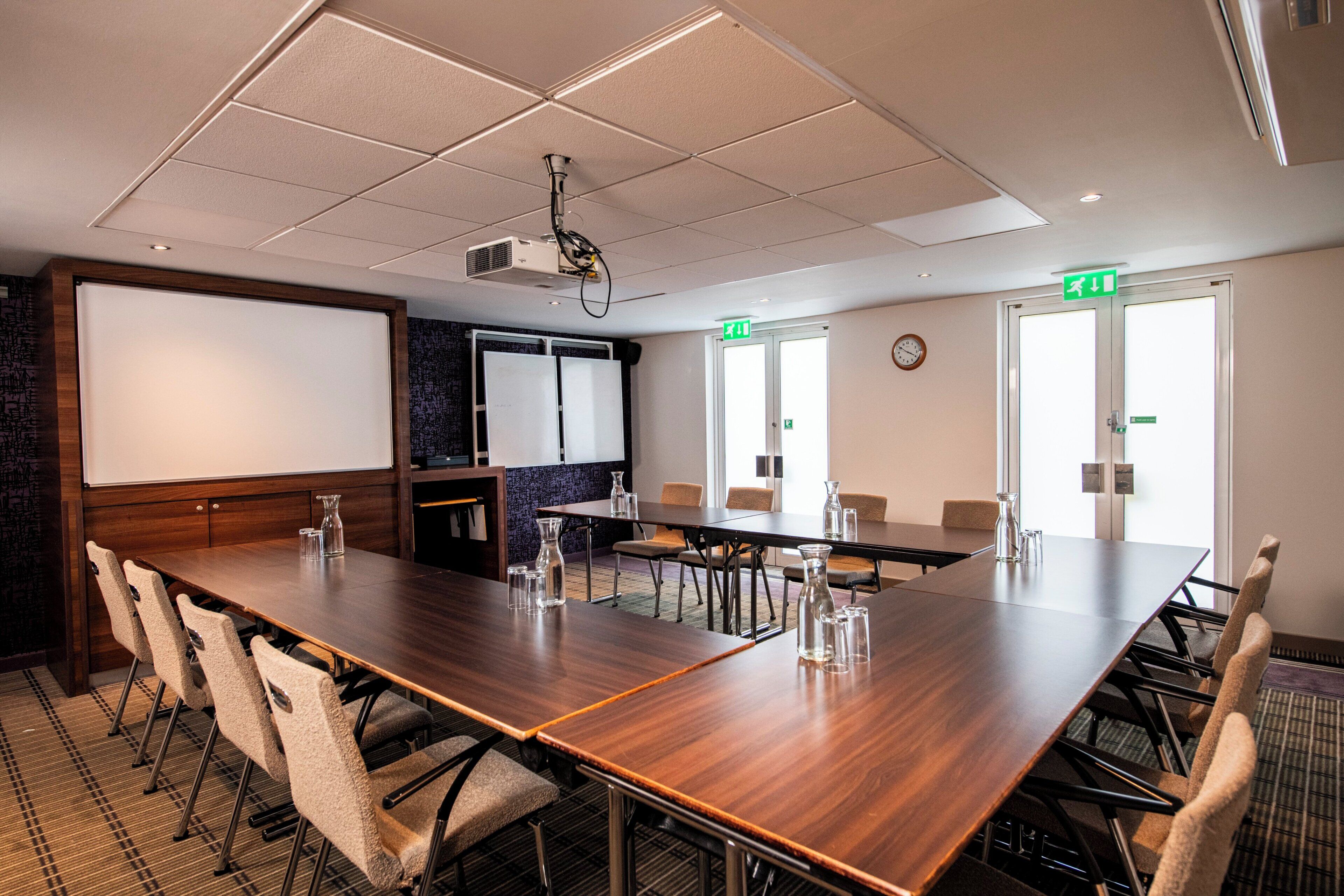 A conference room with a long wooden table, multiple chairs, a projector, a whiteboard, and two exit doors.
