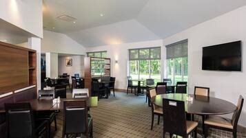 A modern dining area with multiple tables and chairs, large windows with blinds, and a flat-screen TV mounted on the wall.