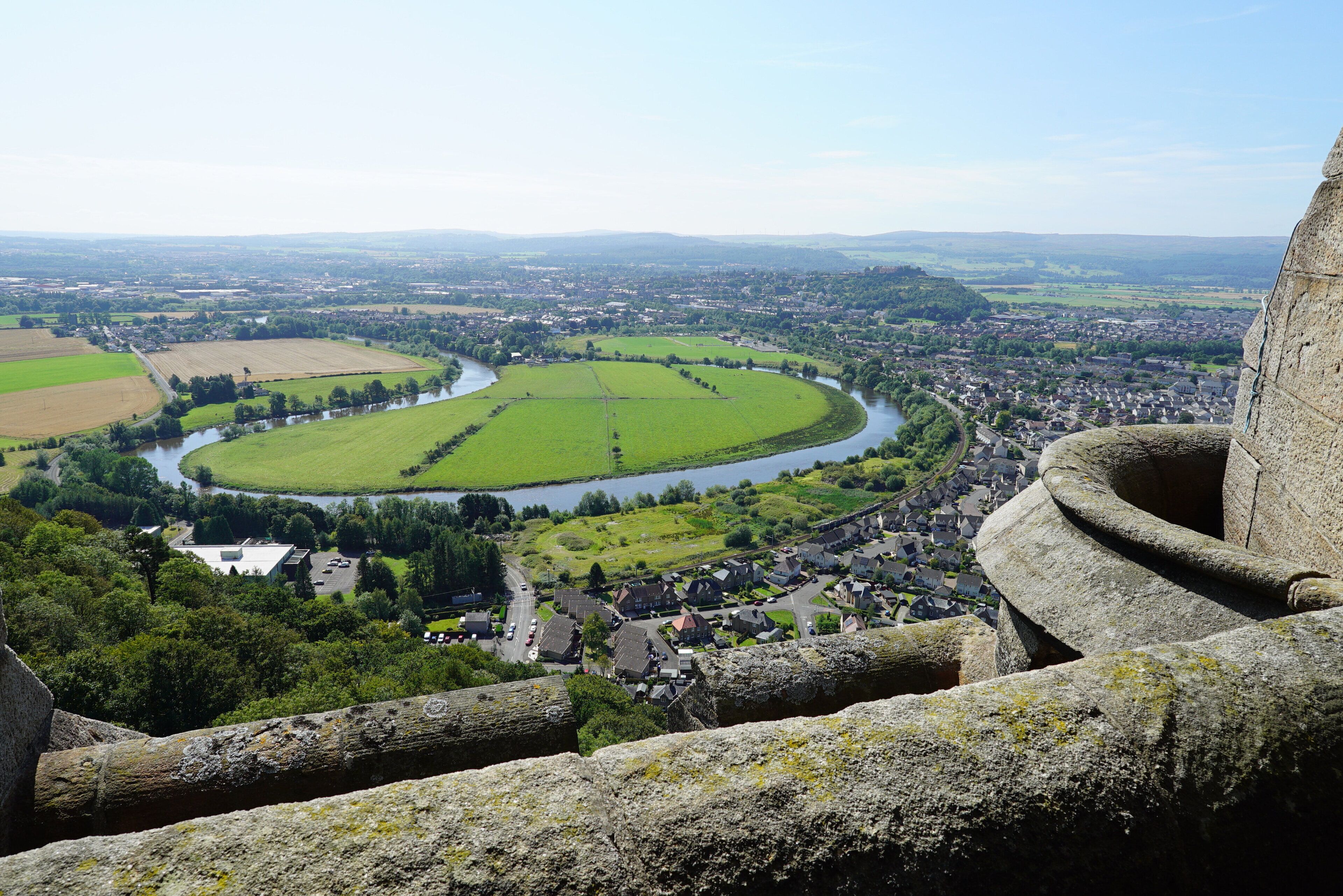 Weitläufige Aussicht auf eine Stadt mit einem Fluss, umgeben von grünen Feldern und einer Steinstruktur im Vordergrund.