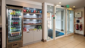 A hotel lobby with a beverage vending machine stocked with various drinks, snack shelves, and a glass door entrance. - Holiday Inn Express Stirling by IHG (Stirling)