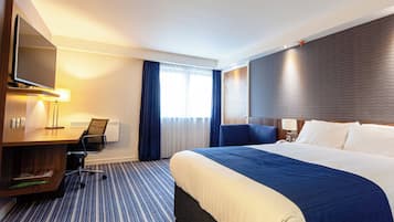 Habitación de hotel con una cama grande, un escritorio con silla, televisión y ventana con cortinas.