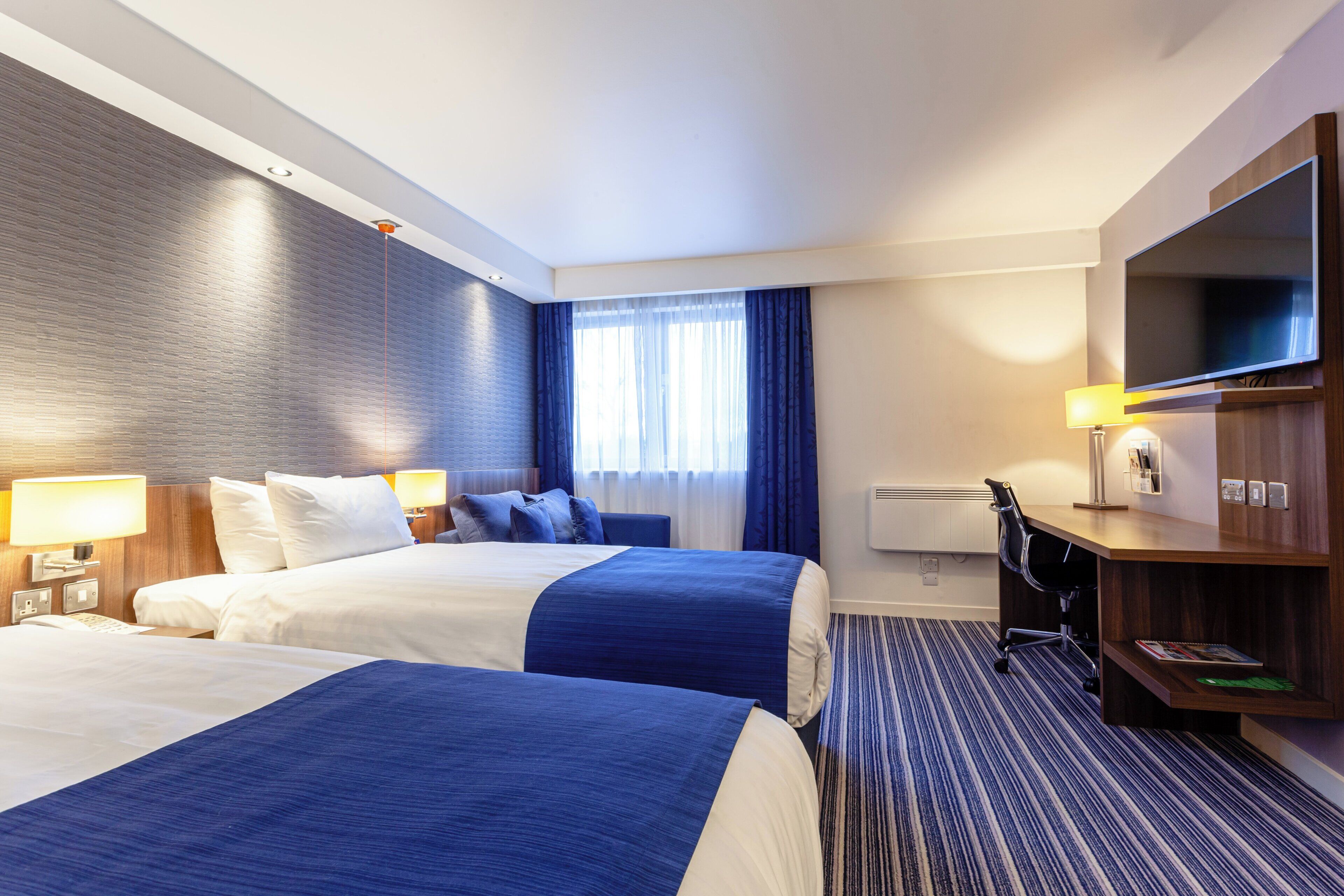 A hotel room with two beds, a desk, a television, and a window with curtains.