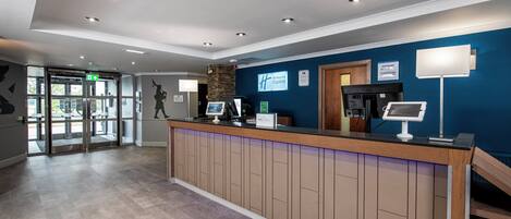 A hotel reception area with a wooden reception desk, a digital screen, and a blue wall.