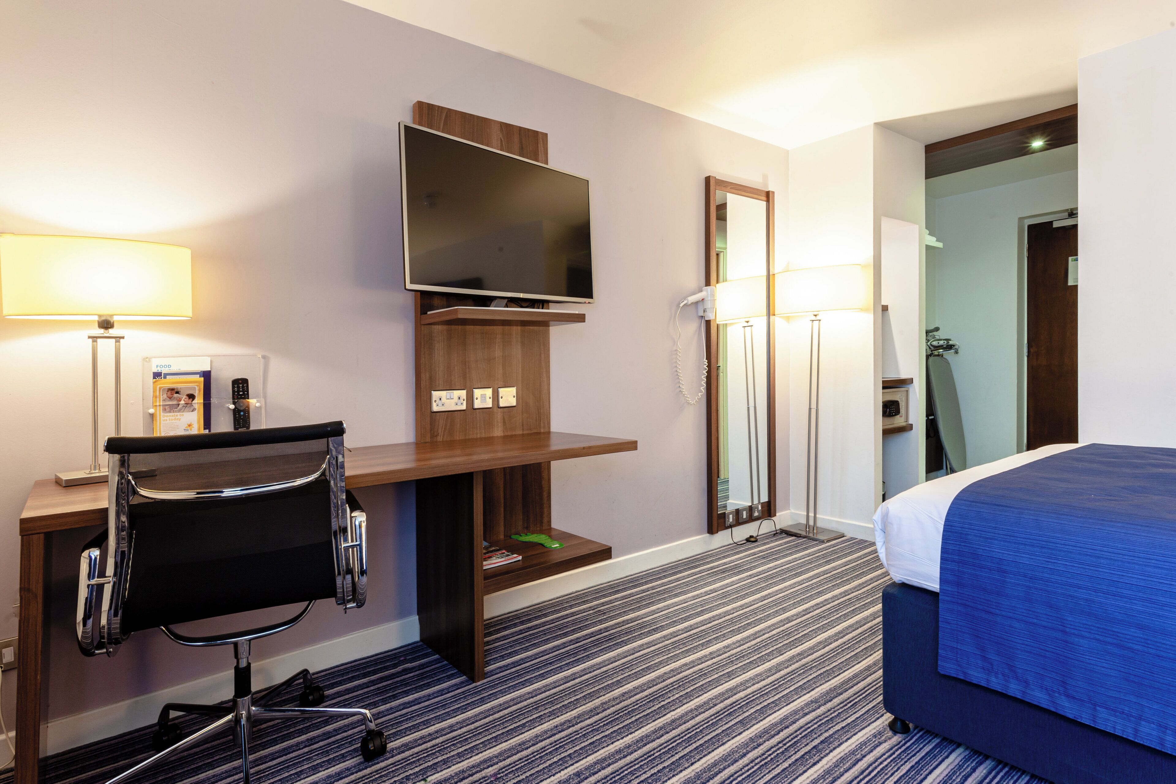 A hotel room with a bed, a desk with a chair, a flat-screen TV, a mirror, and a lamp.