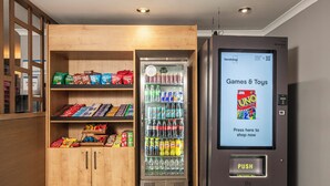 A vending machine labeled "Vendologi" with a digital screen advertising "Games & Toys" and a push button. Adjacent is a wooden cabinet stocked with snacks and beverages. - Holiday Inn Express Edinburgh Airport by IHG (Newbridge)