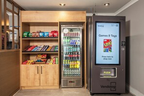 A vending machine labeled "Vendologi" with a digital screen advertising "Games & Toys" and a push button. Adjacent is a wooden cabinet stocked with snacks and beverages. - Holiday Inn Express Edinburgh Airport by IHG (Newbridge)