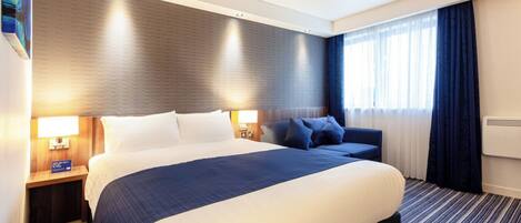 A hotel room with a large bed, two bedside tables with lamps, a sofa, and a window with curtains.