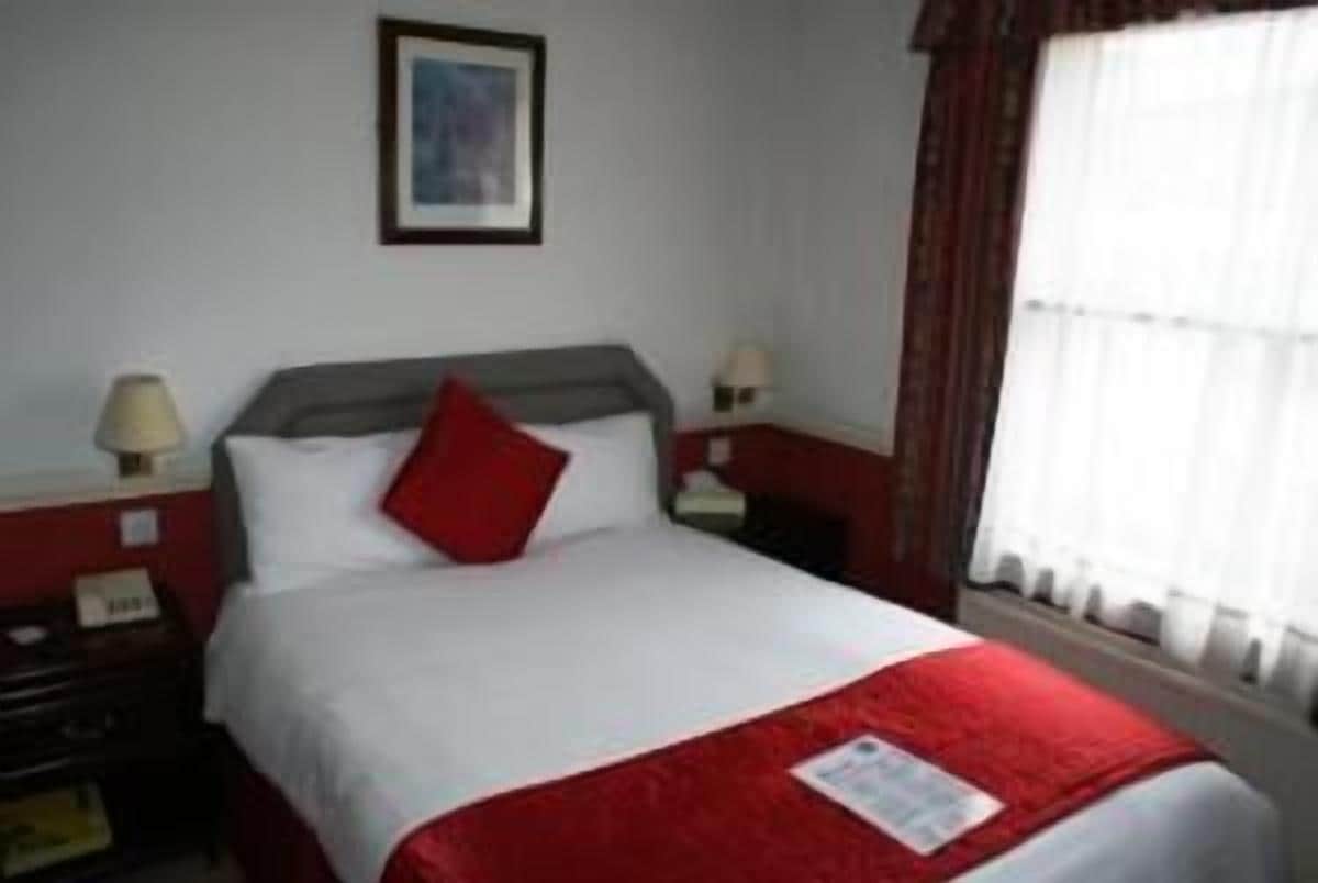 A hotel room with a bed, two bedside tables, a nightstand, a lamp, and a window with curtains.