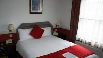 A hotel room with a bed, two bedside tables, a nightstand, a lamp, and a window with curtains.