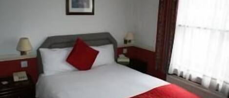 Une chambre d’hôtel comprenant un lit, deux tables de chevet, une petite table, une lampe et une fenêtre avec des rideaux.