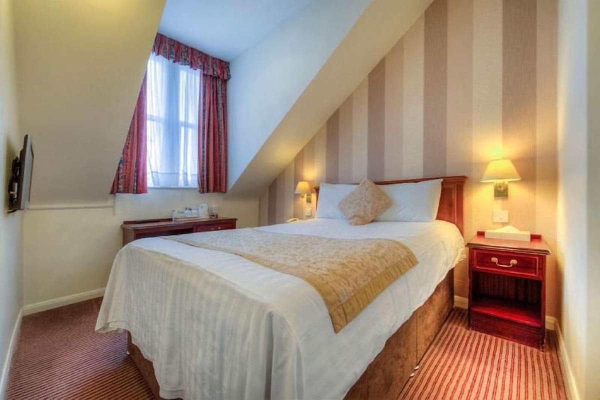 A hotel room with a bed, two bedside tables, a window with curtains, and a striped wall.