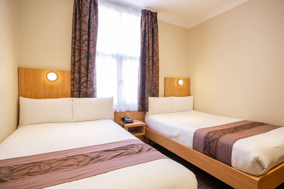 Hotellihuone, jossa on kaksi sänkyä, puinen pääty, yöpöytä lampun kanssa ja ikkuna, jossa on verhot.