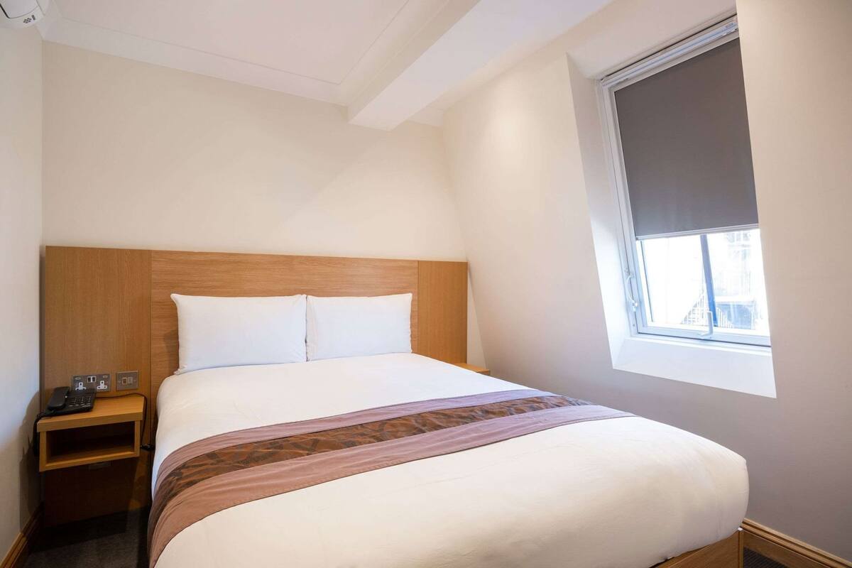 Hotellihuone, jossa on sänky, yöpöytä, jossa on puhelin, ikkuna, jossa on sälekaihtimet, ja näkymä ulos.